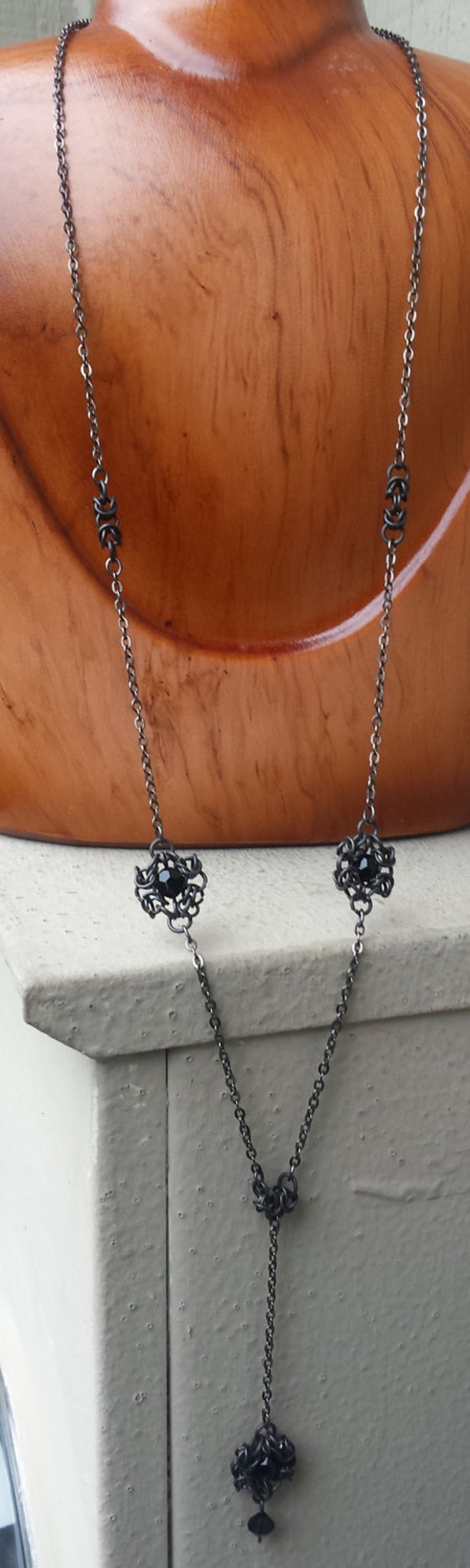 May include: A black chain necklace with three black gemstone accents in a delicate, intricate design. The necklace is displayed on a wooden mannequin.