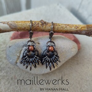May include: A pair of black metal earrings with a copper bead accent. The earrings are in the shape of a fan and have a delicate, intricate design. The earrings are hanging from a wooden branch.