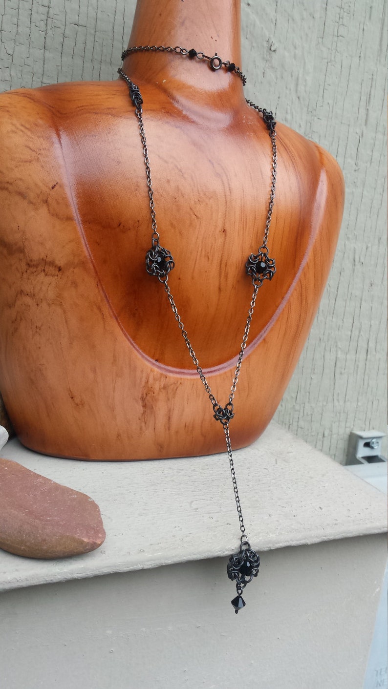 May include: A long black chain necklace with four black metal ball charms. The necklace is displayed on a wooden mannequin.