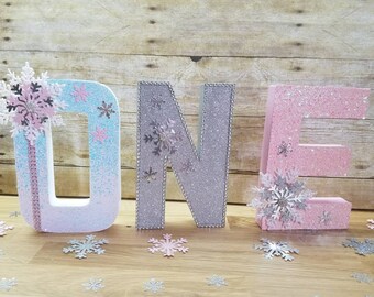 Könnte beinhalten: Glitzernde Buchstaben "ONE" in Pastellfarben, verziert mit Schneeflocken. Das "O" ist hellblau und rosa, das "N" ist silber und das "E" ist rosa. Dekorative Schneeflocken sind verstreut und erzeugen eine festliche Atmosphäre.