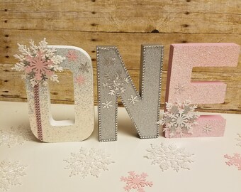 Könnte beinhalten: Dekorative Buchstaben, die "ONE" für einen ersten Geburtstag buchstabieren. Das "O" ist weiß mit rosa und silbernen Akzenten, das "N" ist silber und das "E" ist rosa. Weiße und rosa Schneeflocken-Dekorationen sind verstreut.