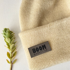 May include: Cream-colored knit beanie with a rectangular gray leather tag. The tag features the text "BOOM MARKETING" in a dark gray font. The beanie is made of a soft, textured material.