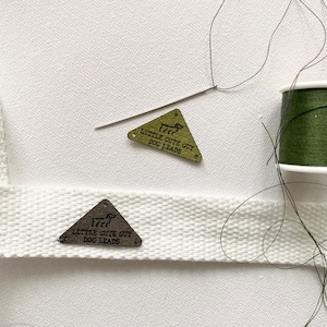 May include: Two metal triangle-shaped dog leash tags with the text "Little Cute Guy Dog Leads" on a white background. One tag is gold and the other is silver.