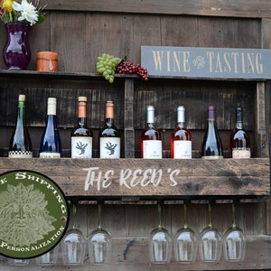 May include: A dark brown wooden wall-mounted wine rack featuring a shelf for eight wine bottles and a lower rack for hanging wine glasses. The front panel is engraved with the text THE REED'S, and a sign above reads WINE TASTING.