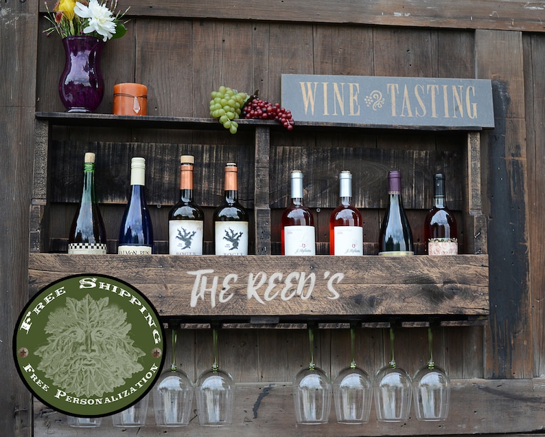 May include: A dark brown wooden wall-mounted wine rack featuring a shelf for eight wine bottles and a lower rack for hanging wine glasses. The front panel is engraved with the text THE REED'S, and a sign above reads WINE TASTING.