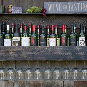 May include: A dark-stained wooden wall-mounted wine rack featuring a top shelf with various wine bottles, green and red grapes, and a sign that reads WINE TASTING. Below the shelf, ten clear glass wine glasses hang upside down by their stems.