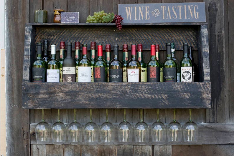 May include: A dark-stained wooden wall-mounted wine rack featuring a top shelf with various wine bottles, green and red grapes, and a sign that reads WINE TASTING. Below the shelf, ten clear glass wine glasses hang upside down by their stems.