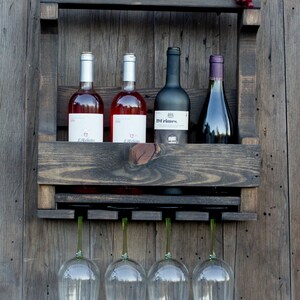 May include: A dark stained wooden wall-mounted wine rack featuring a top shelf with a red candle, a small brown ceramic pot, a sign reading Family Life's Greatest Blessing, and faux grapes. It holds four wine bottles and four hanging glass stems.