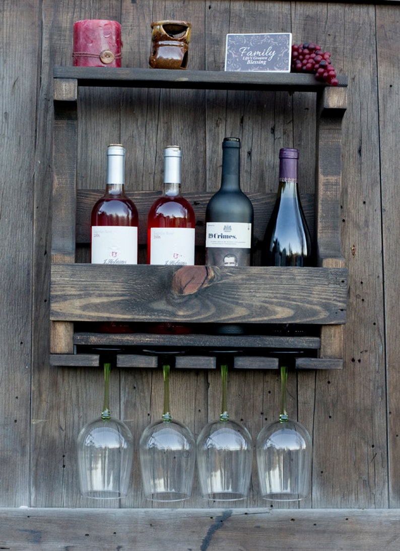 May include: A dark stained wooden wall-mounted wine rack featuring a top shelf with a red candle, a small brown ceramic pot, a sign reading Family Life's Greatest Blessing, and faux grapes. It holds four wine bottles and four hanging glass stems.