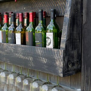 May include: A wall-mounted black wooden wine rack with a distressed finish, holding multiple glass wine bottles on the top shelf and featuring a row of hanging clear glass wine stems underneath, with a sign reading WINE TASTING above.