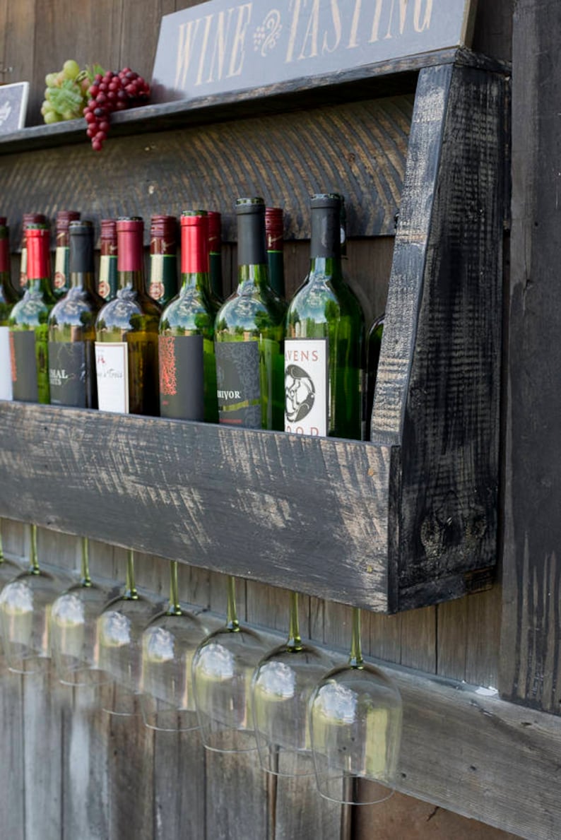 May include: A wall-mounted black wooden wine rack with a distressed finish, holding multiple glass wine bottles on the top shelf and featuring a row of hanging clear glass wine stems underneath, with a sign reading WINE TASTING above.
