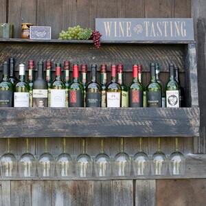 May include: A dark stained wooden wall-mounted wine rack holding sixteen glass wine bottles with various labels. The rack features a top shelf displaying green and red grapes, a small sign reading WINE TASTING, and nine wine glasses hanging below.