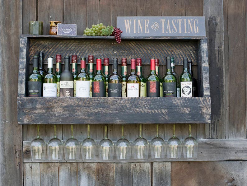 May include: A dark stained wooden wall-mounted wine rack holding sixteen glass wine bottles with various labels. The rack features a top shelf displaying green and red grapes, a small sign reading WINE TASTING, and nine wine glasses hanging below.
