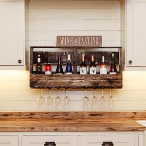 May include: A dark brown wooden wall-mounted wine rack featuring a horizontal shelf for eight wine bottles and a lower rack for hanging eight wine glasses. A rectangular sign with the text WINE TASTING is positioned on top of the wooden rack.