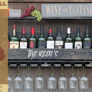 May include: A black wooden wall-mounted wine rack featuring a shelf for bottles, a stemware rack for hanging glasses, and the text "THE REED'S" painted on the front. The rack holds various wine bottles and is topped with red and green grapes.
