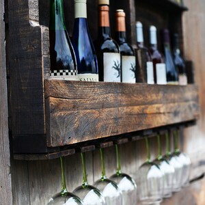 May include: A dark stained wooden wall-mounted wine rack featuring a shelf for multiple wine bottles and an integrated rack underneath for hanging wine glasses. The rack is filled with various wine bottles and clear glass stemware.