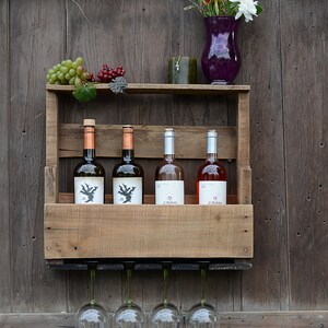 May include: A wall-mounted wooden wine rack featuring a top shelf holding green grapes, a dark green candle, and a purple glass vase with yellow and white flowers. Four wine bottles sit on the middle shelf, and four wine glasses hang below.
