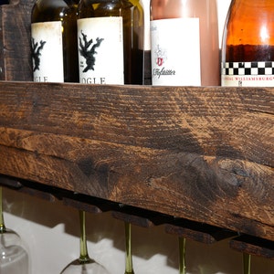 May include: A dark brown stained wooden wall-mounted wine rack featuring a top shelf for storing multiple wine bottles and a lower section with integrated slots designed to hold and display several stemmed wine glasses hanging upside down.