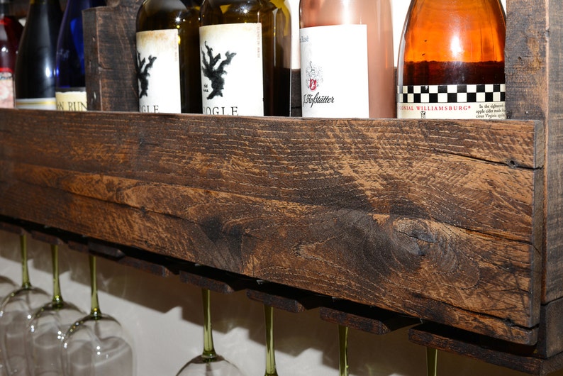 May include: A dark brown stained wooden wall-mounted wine rack featuring a top shelf for storing multiple wine bottles and a lower section with integrated slots designed to hold and display several stemmed wine glasses hanging upside down.