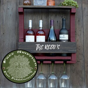 May include: A wall-mounted wooden wine rack featuring a dark stained shelf and deep red side supports. The front panel is engraved with the text THE REED'S. It holds four wine bottles and includes a rack underneath for hanging three wine glasses.