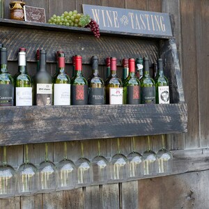 May include: A dark stained wooden wall-mounted wine rack featuring a shelf holding eleven assorted wine bottles and a top ledge with grapes and a sign reading WINE TASTING. Below the shelf, nine clear glass wine glasses hang upside down.
