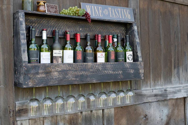 May include: A dark stained wooden wall-mounted wine rack featuring a shelf holding eleven assorted wine bottles and a top ledge with grapes and a sign reading WINE TASTING. Below the shelf, nine clear glass wine glasses hang upside down.