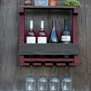 May include: A wall-mounted wooden wine rack featuring a dark brown shelf and a deep red frame. It holds four wine bottles and includes a rack underneath for four hanging wine glasses. The top shelf displays a small sign reading Family Life's Greatest Blessing.