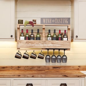 May include: A light brown wooden wall-mounted rack featuring two shelves for eight wine bottles, a top sign reading WINE TASTING, and a front panel with the text You'll be my Glass of Wine, I'll be your Shot of Whiskey, with four mugs and four wine glasses.