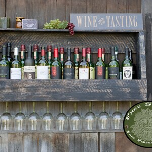 May include: A dark-stained wooden wall-mounted wine rack featuring a top shelf holding multiple wine bottles, green and red grapes, and a sign that reads WINE TASTING. Below the shelf, eight clear glass wine glasses hang upside down.