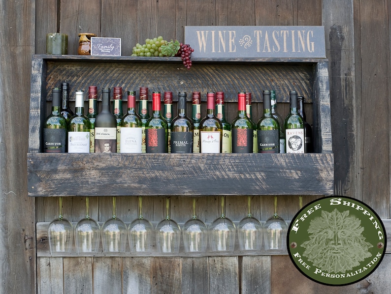 May include: A dark-stained wooden wall-mounted wine rack featuring a top shelf holding multiple wine bottles, green and red grapes, and a sign that reads WINE TASTING. Below the shelf, eight clear glass wine glasses hang upside down.