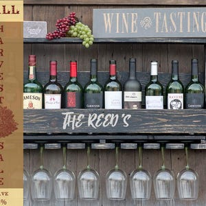 May include: A dark wood wall-mounted wine rack featuring the text THE REED'S, holding eight wine bottles and seven hanging wine glasses. Above the rack, a sign reads WINE TASTING, with clusters of red and green grapes resting on the top shelf.