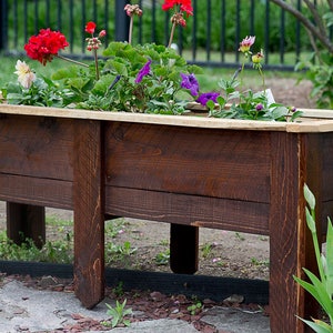 May include: A rectangular, dark brown wooden planter box, filled with colourful flowers. The flowers include red geraniums, purple petunias, and white and pink blooms. The planter has four legs and is suitable for a garden or patio.