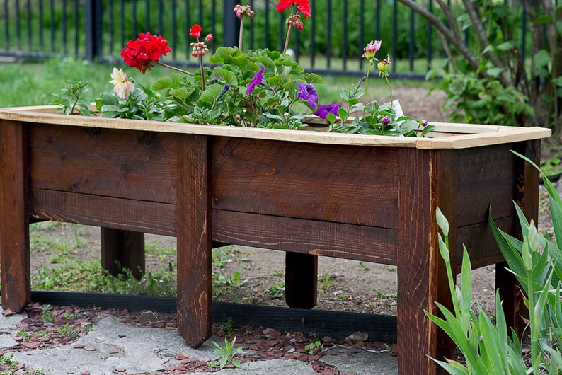 May include: A rectangular, dark brown wooden planter box, filled with colourful flowers. The flowers include red geraniums, purple petunias, and white and pink blooms. The planter has four legs and is suitable for a garden or patio.