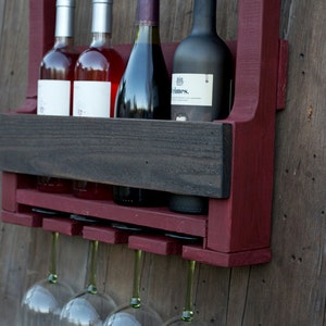May include: A wall-mounted wine rack crafted from dark red and charcoal-stained wood, featuring a top shelf, a middle section holding four wine bottles, and a bottom rack with four clear glass wine glasses hanging upside down by their stems.