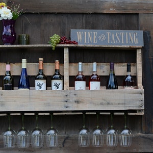 May include: A wooden pallet wine rack featuring a shelf holding eight wine bottles and a sign that reads WINE TASTING. Below the shelf, eight clear glass wine glasses hang upside down. A purple vase with white flowers sits on the top shelf.
