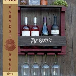 May include: A wall-mounted wooden wine rack with a dark stained finish and maroon accents, featuring the text "THE REED'S" on the front. It holds four wine bottles and has a rack underneath for hanging four wine glasses. A vertical banner reads "FALL HARVEST SALE SAVE 15%" with a green man icon.