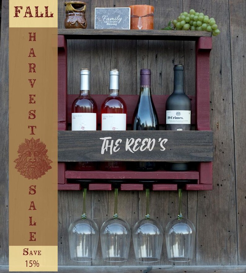 May include: A wall-mounted wooden wine rack with a dark stained finish and maroon accents, featuring the text "THE REED'S" on the front. It holds four wine bottles and has a rack underneath for hanging four wine glasses. A vertical banner reads "FALL HARVEST SALE SAVE 15%" with a green man icon.
