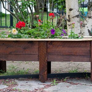 May include: A rectangular wooden planter box filled with colourful flowers. The planter is made of dark brown wood and has a light-coloured wooden trim around the top. The flowers include red, purple, and yellow blooms.