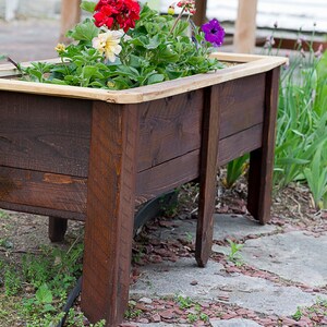 May include: A dark brown, rectangular wooden planter box filled with colourful flowers. The planter has a light wood trim and stands on four legs. The flowers include red, yellow, and purple blooms. The planter is suitable for a garden.