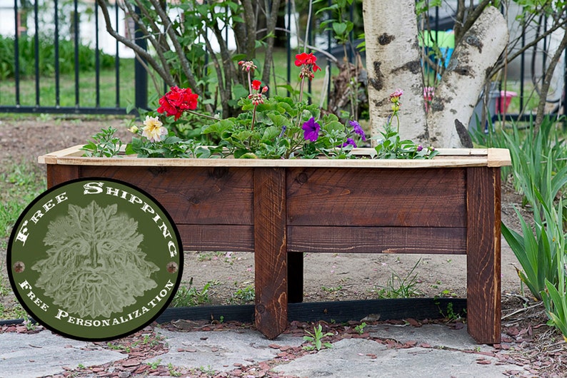May include: A dark stained wooden planter box filled with vibrant flowers. The box has a natural wood trim and is rectangular. The flowers include red, purple, and yellow blooms. The planter is outdoors in a garden.