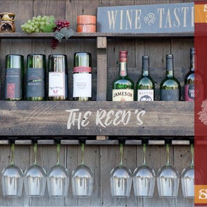 May include: A dark brown wooden wall-mounted wine rack featuring two shelves, eight glass holders, and the text "THE REED'S" engraved on the front. The top shelf holds a small ceramic oil burner, green and red grapes, and a candle. Eight wine bottles are displayed.