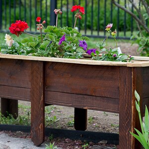 May include: A dark brown wooden planter box filled with vibrant flowers. The box has a light wood trim and is supported by sturdy legs. The flowers include red, purple, and white blooms, adding a splash of colour.