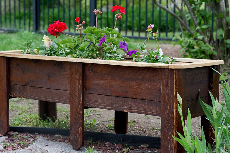 May include: A dark brown wooden planter box filled with vibrant flowers. The box has a light wood trim and is supported by sturdy legs. The flowers include red, purple, and white blooms, adding a splash of colour.