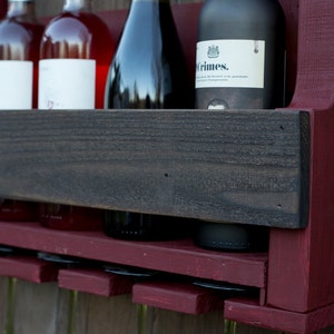 May include: A wall-mounted wine rack crafted from dark red stained wood, featuring a horizontal black wooden shelf for bottle storage and a lower section with slots for hanging wine glasses. Four wine bottles are positioned on the shelf.