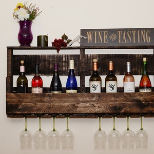 May include: A dark brown wooden wall-mounted wine rack featuring a shelf for eight wine bottles and a lower rack for eight hanging wine glasses. A sign reading WINE TASTING sits on top, alongside a purple vase with flowers and a corkscrew.
