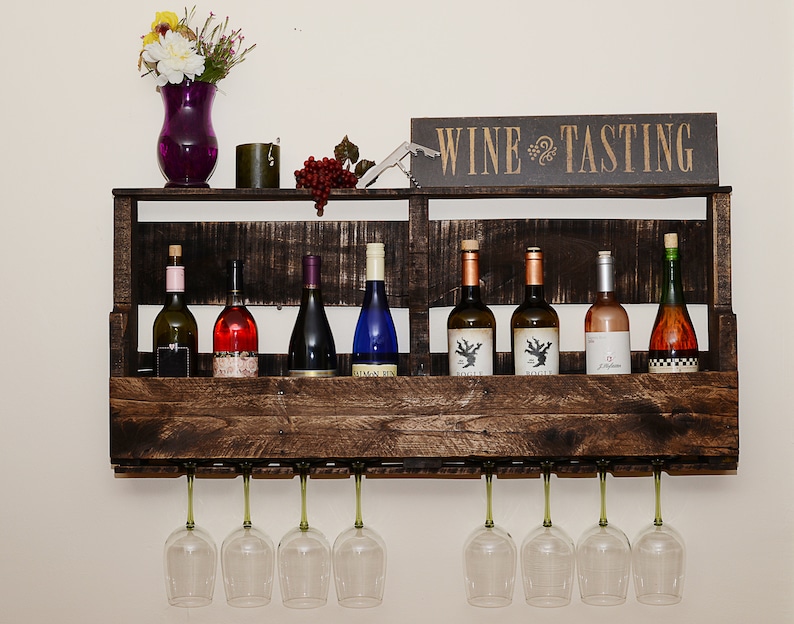 May include: A dark brown wooden wall-mounted wine rack featuring a shelf for eight wine bottles and a lower rack for eight hanging wine glasses. A sign reading WINE TASTING sits on top, alongside a purple vase with flowers and a corkscrew.