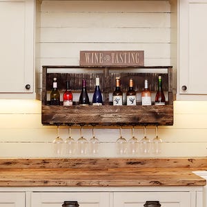 May include: A dark brown wooden wall-mounted wine rack featuring a horizontal shelf for eight wine bottles and a lower rack for hanging eight wine glasses. A rectangular sign with the text WINE TASTING is positioned on top of the wooden rack.