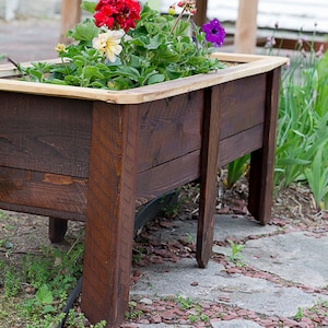 May include: A dark brown wooden raised garden bed, filled with colourful flowers. The planter has a natural wood trim. The flowers include red, yellow, and purple blooms, creating a vibrant display. The planter is suitable for a garden.
