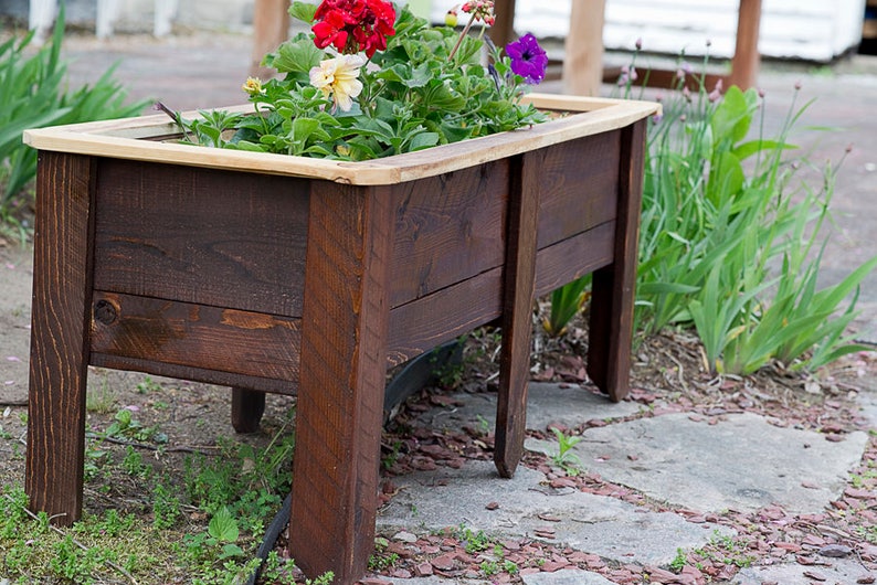 May include: A dark brown wooden raised garden bed, filled with colourful flowers. The planter has a natural wood trim. The flowers include red, yellow, and purple blooms, creating a vibrant display. The planter is suitable for a garden.
