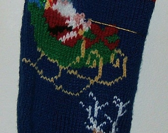Op de afbeelding: Een blauwe gebreide kerstsok met een witte boord en rode bies. De sok heeft een groene en gouden slee met de kerstman en een rendier, en de naam "LUKE" is geborduurd op de boord.